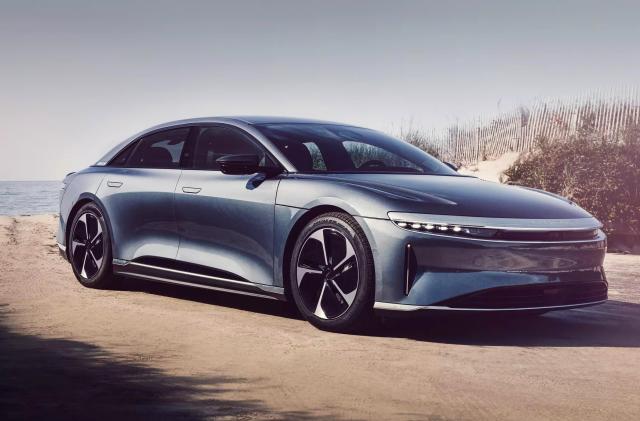 A grey Lucid Air Pure RWD EV is parked among sand dunes on the edge of the beach.